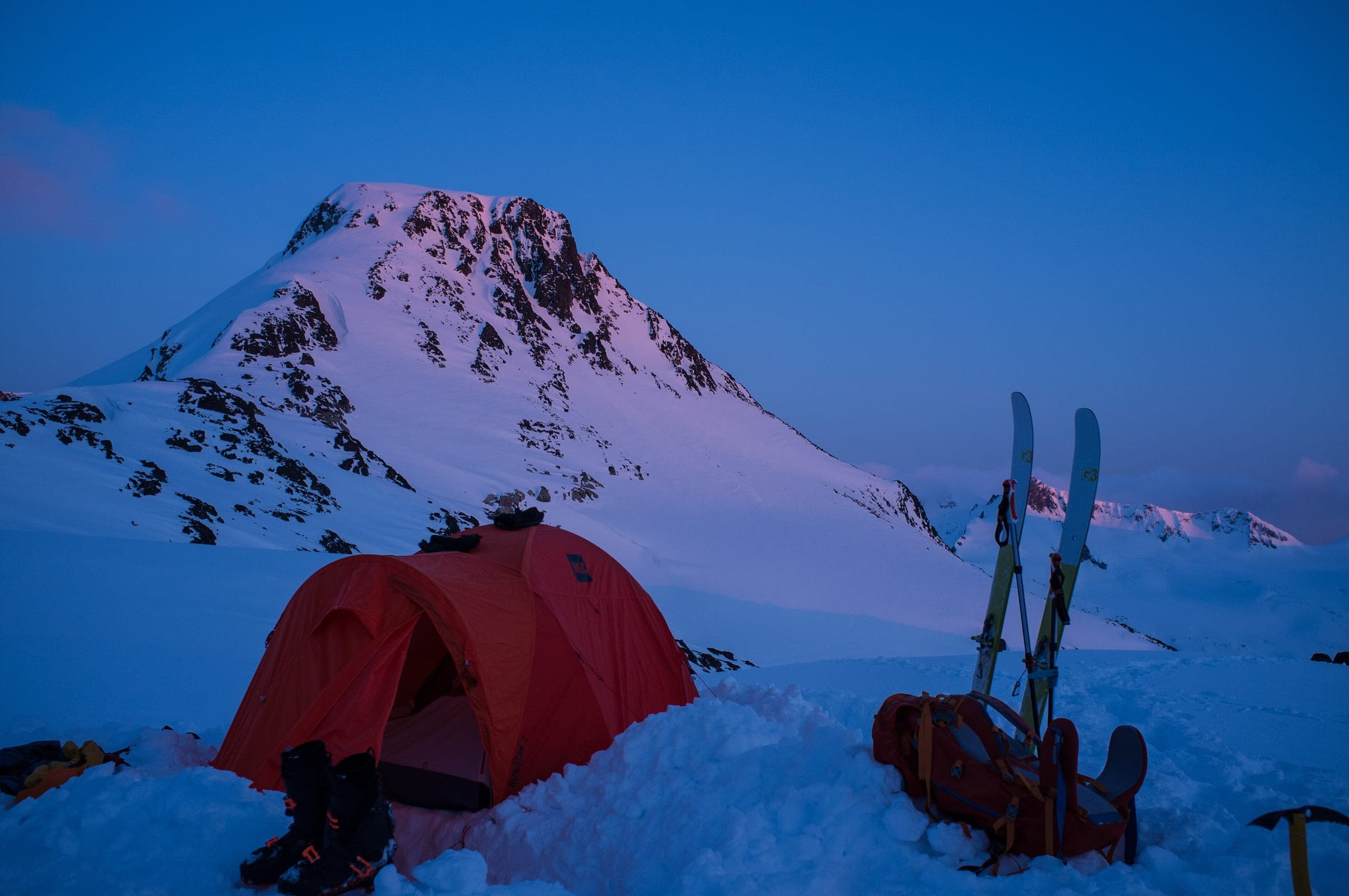 A tent and a pair of touring skis sit in the snow below a peak lit pink at dusk on the Spearhead.
