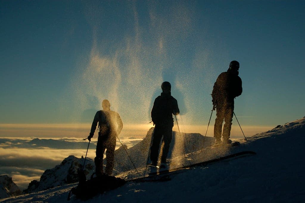 Three skiers stand in blowing snow lit by low sun in the Blackcomb backcountry above the clouds.