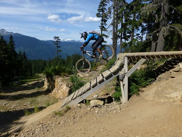 A mountain biker rolls off a wooden ramp in the Whistler Mountain Bike Park with snowy peaks across the valley.
