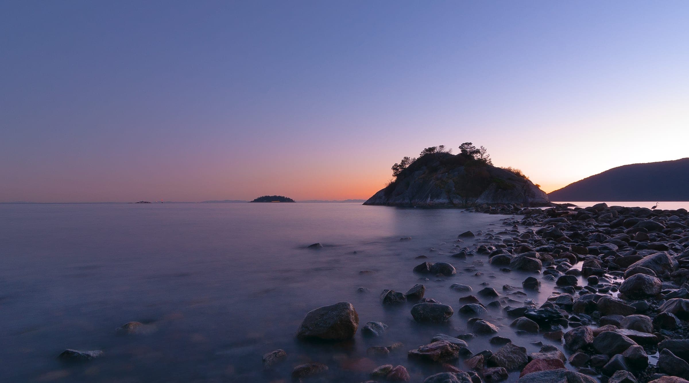 Whytecliff Islet stands dark against an orange dusk sky beyond the rocky beach at Whytecliff Park.