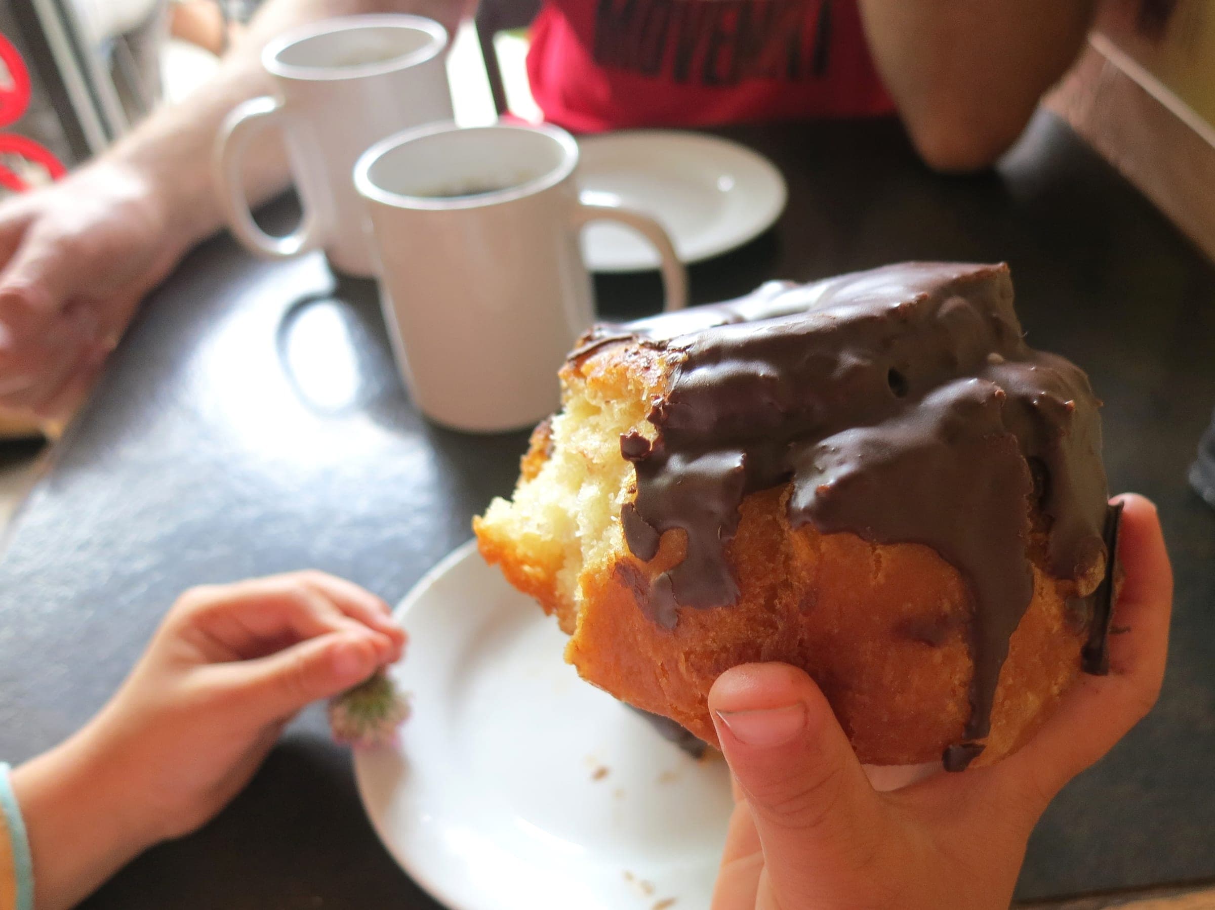 A hand holds a bitten chocolate-glazed doughnut over coffee mugs at Honey Doughnuts in Deep Cove.