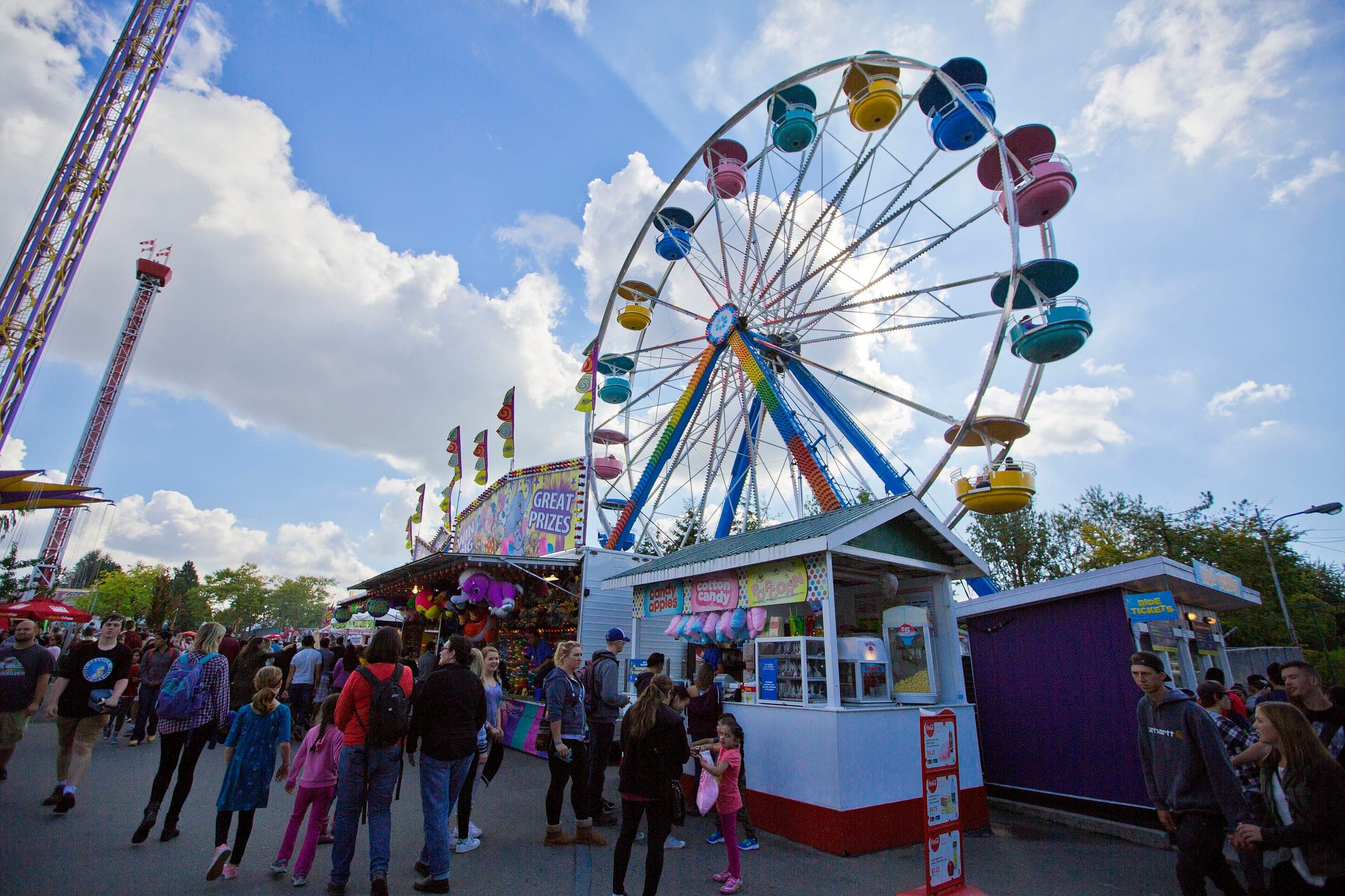 Fairgoers walk the PNE midway at Hastings Park below a Ferris wheel and two drop towers.