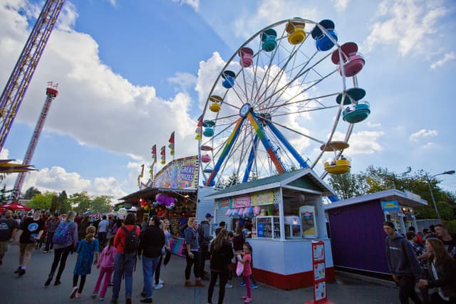 Fairgoers walk the PNE midway at Hastings Park below a Ferris wheel and two drop towers.
