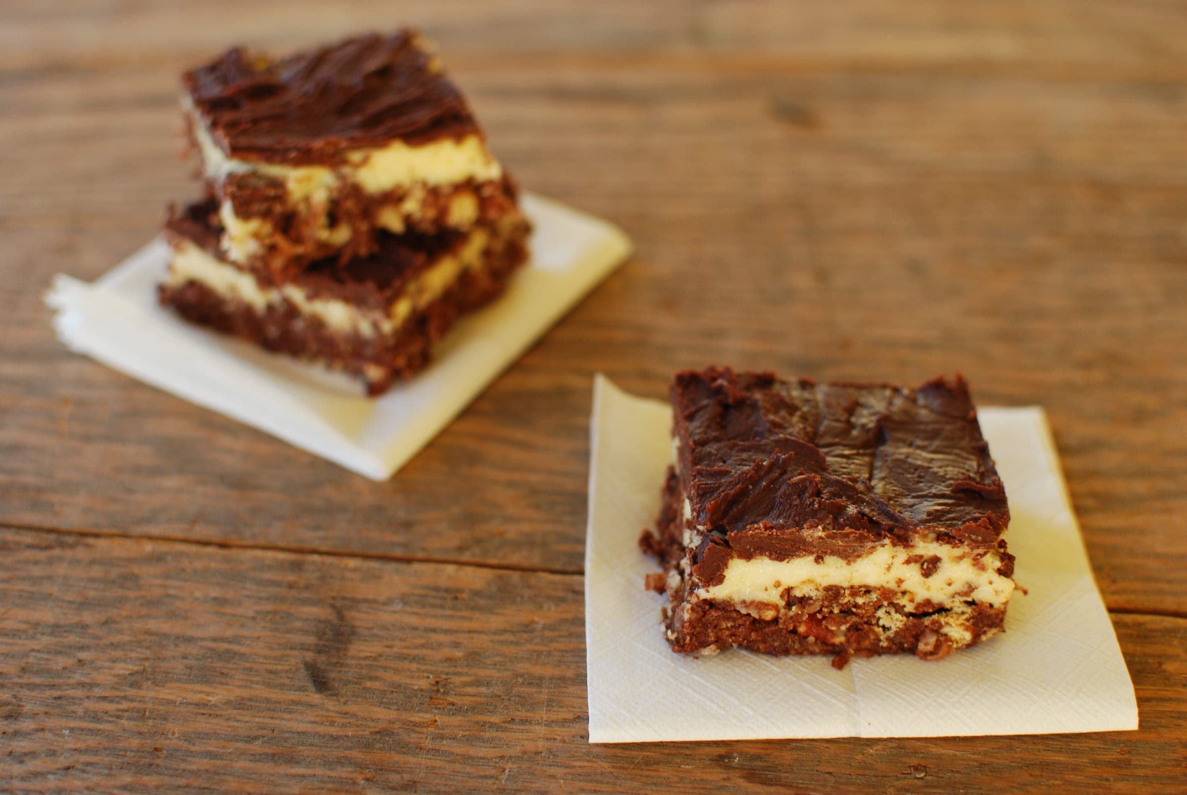Nanaimo bars with a crumb base, custard filling and chocolate top sit on napkins on a wooden table.