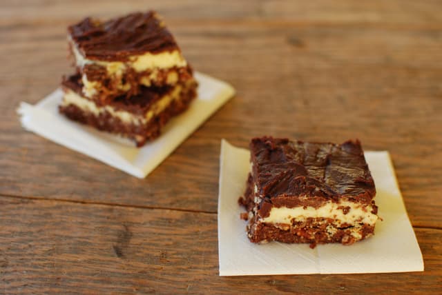 Nanaimo bars with a crumb base, custard filling and chocolate top sit on napkins on a wooden table.