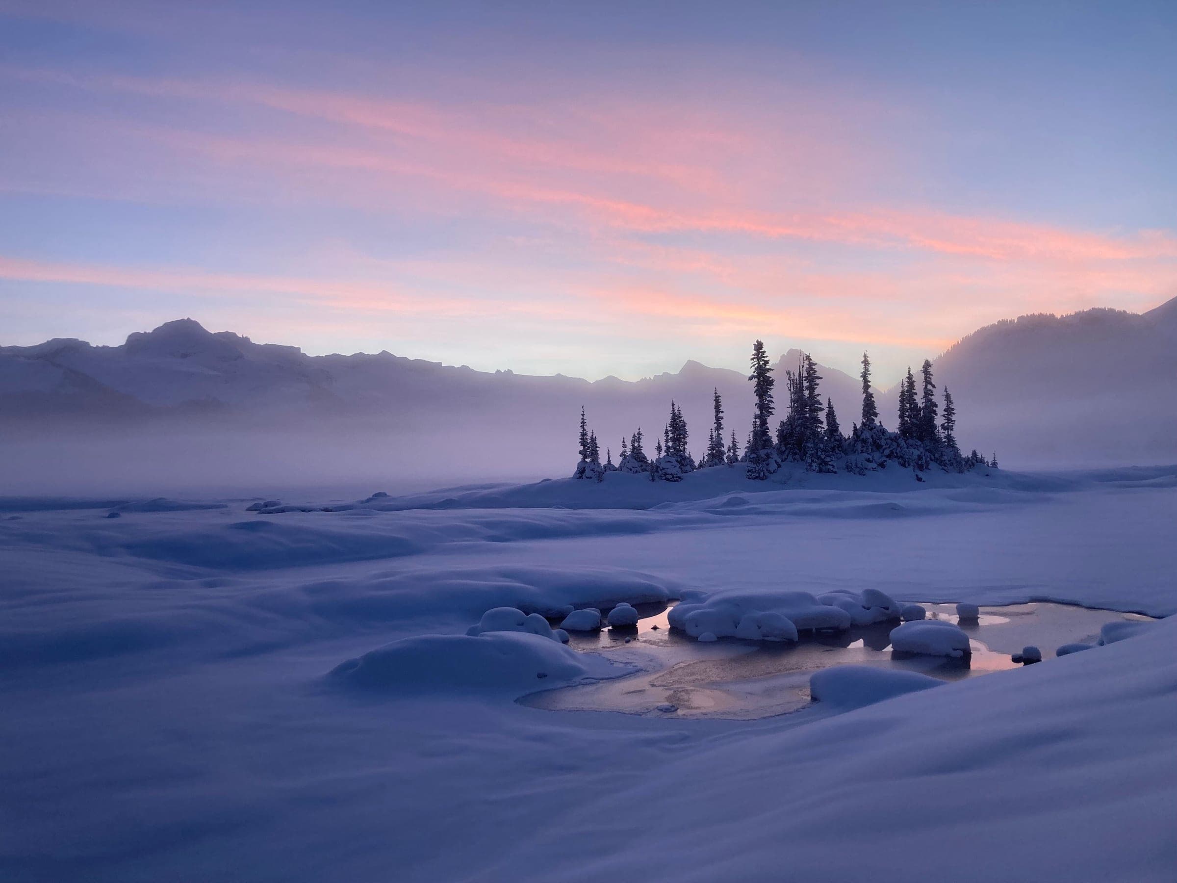 Pink dawn light rises over frozen, snow-covered Garibaldi Lake and its small treed island on New Year's Day.