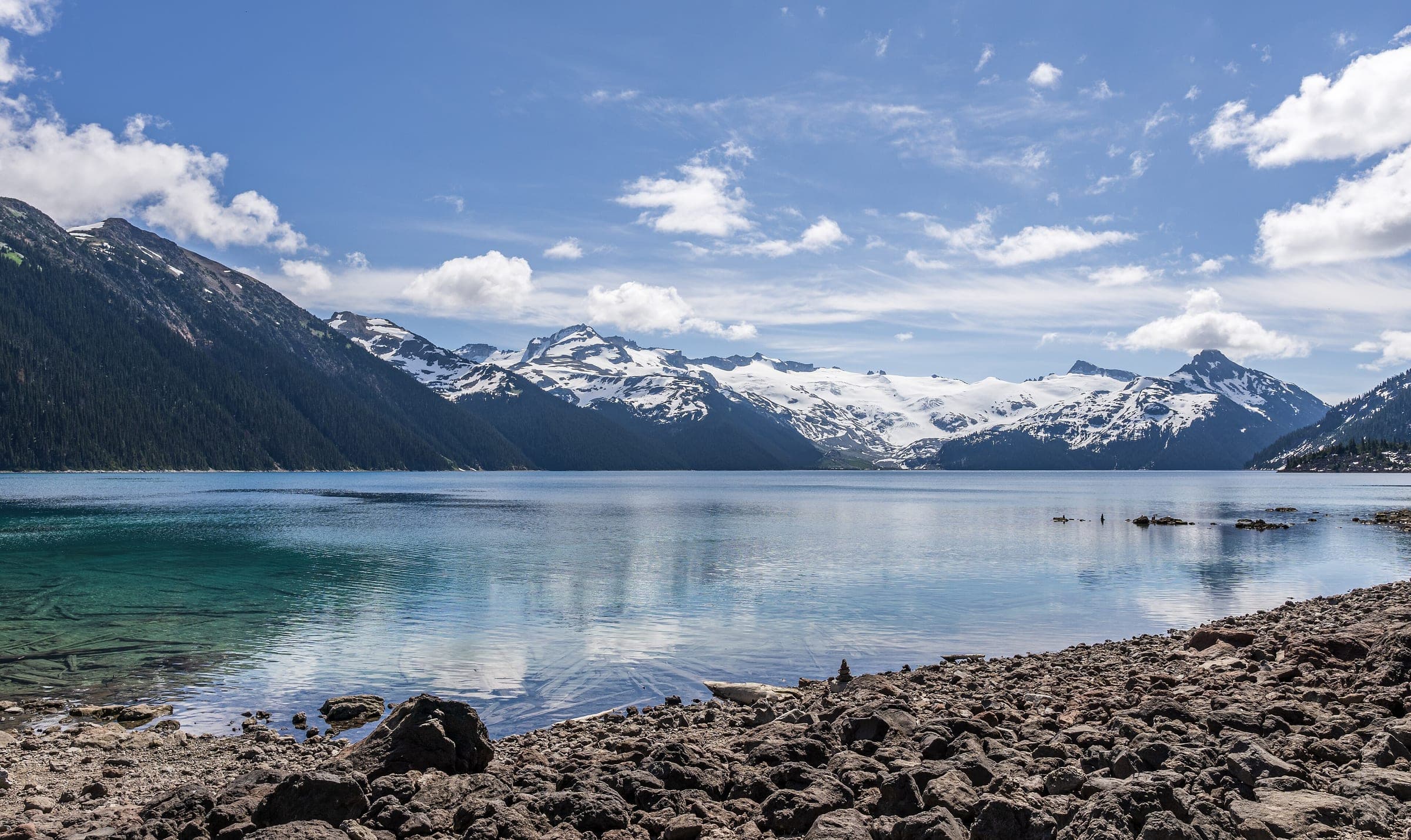 Snowy peaks rise beyond clear water off the rocky shore of Garibaldi Lake in Garibaldi Provincial Park.