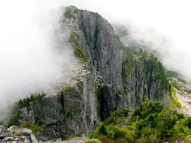 Fog wraps the sheer granite of the West Lion as hikers stand on the ledge below its summit.