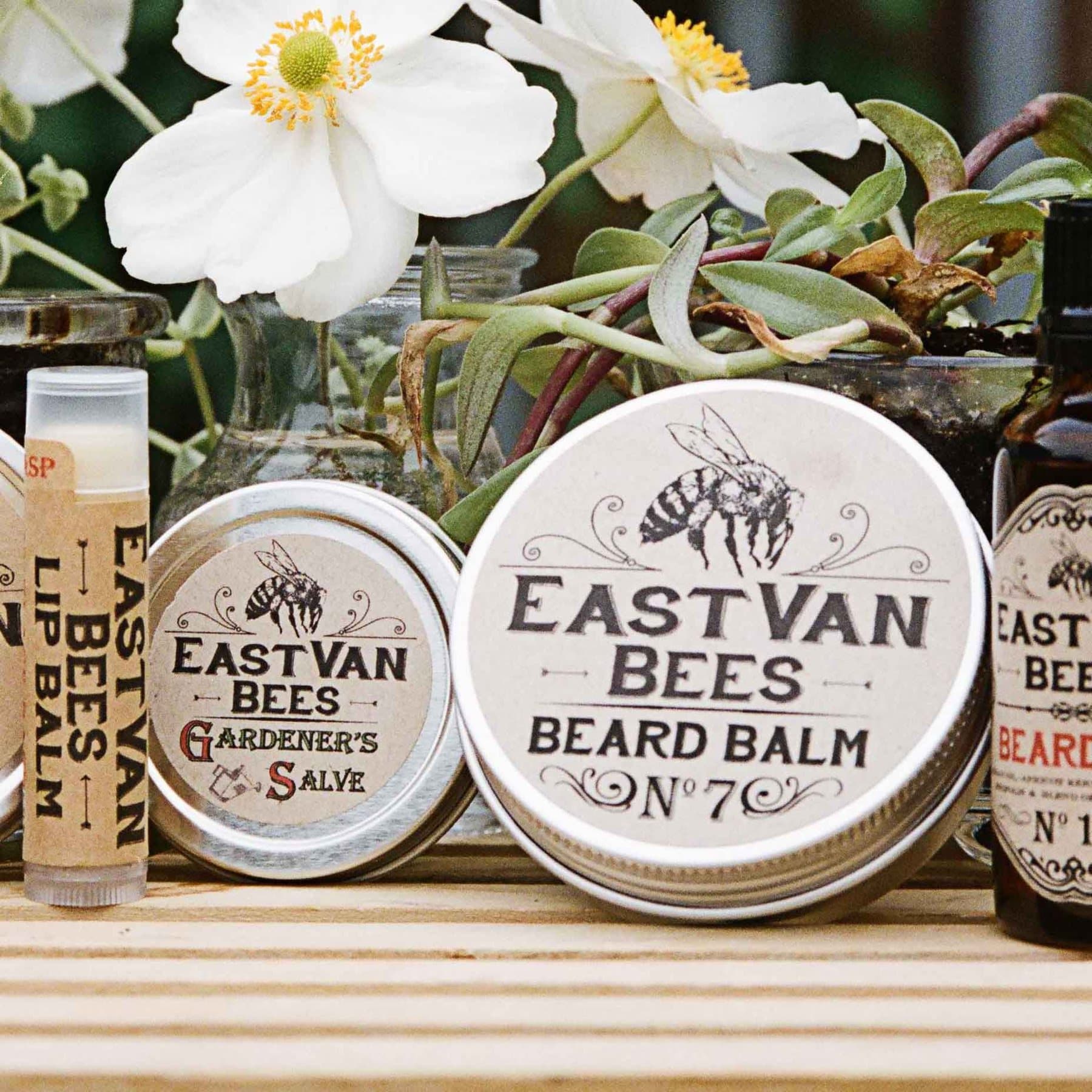 EastVan Bees beard balm, gardener's salve and lip balm set among white flowers on a wooden bench.