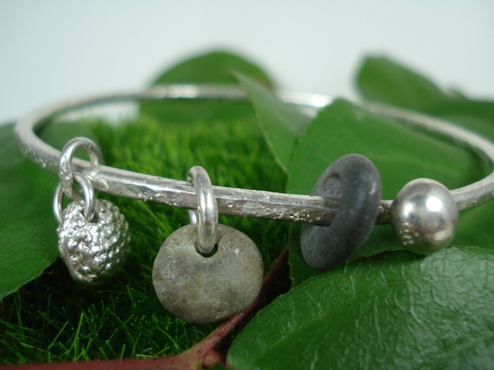 An Effie Baker silver bangle hung with beach pebbles and silver charms, resting on green leaves.