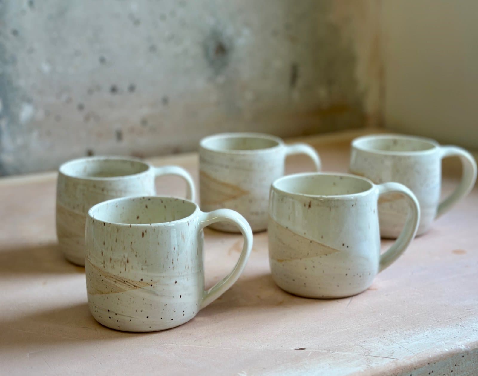 Five speckled JAM Pottery mugs on a pale wooden ledge.