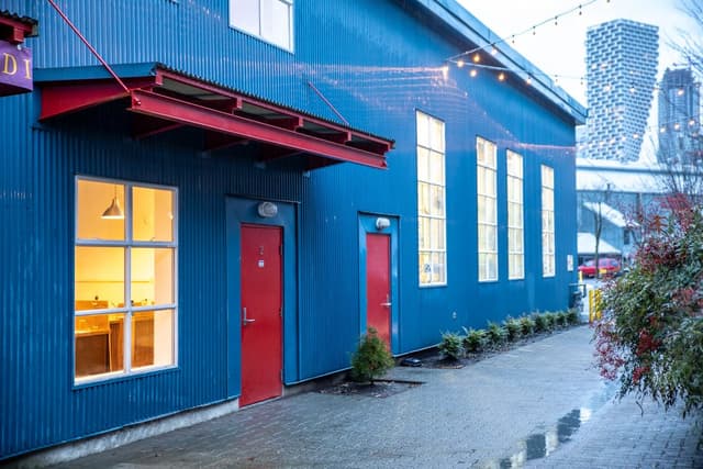 The blue corrugated front of Kasama Chocolate's workshop and store on Granville Island, with red doors and lit windows.