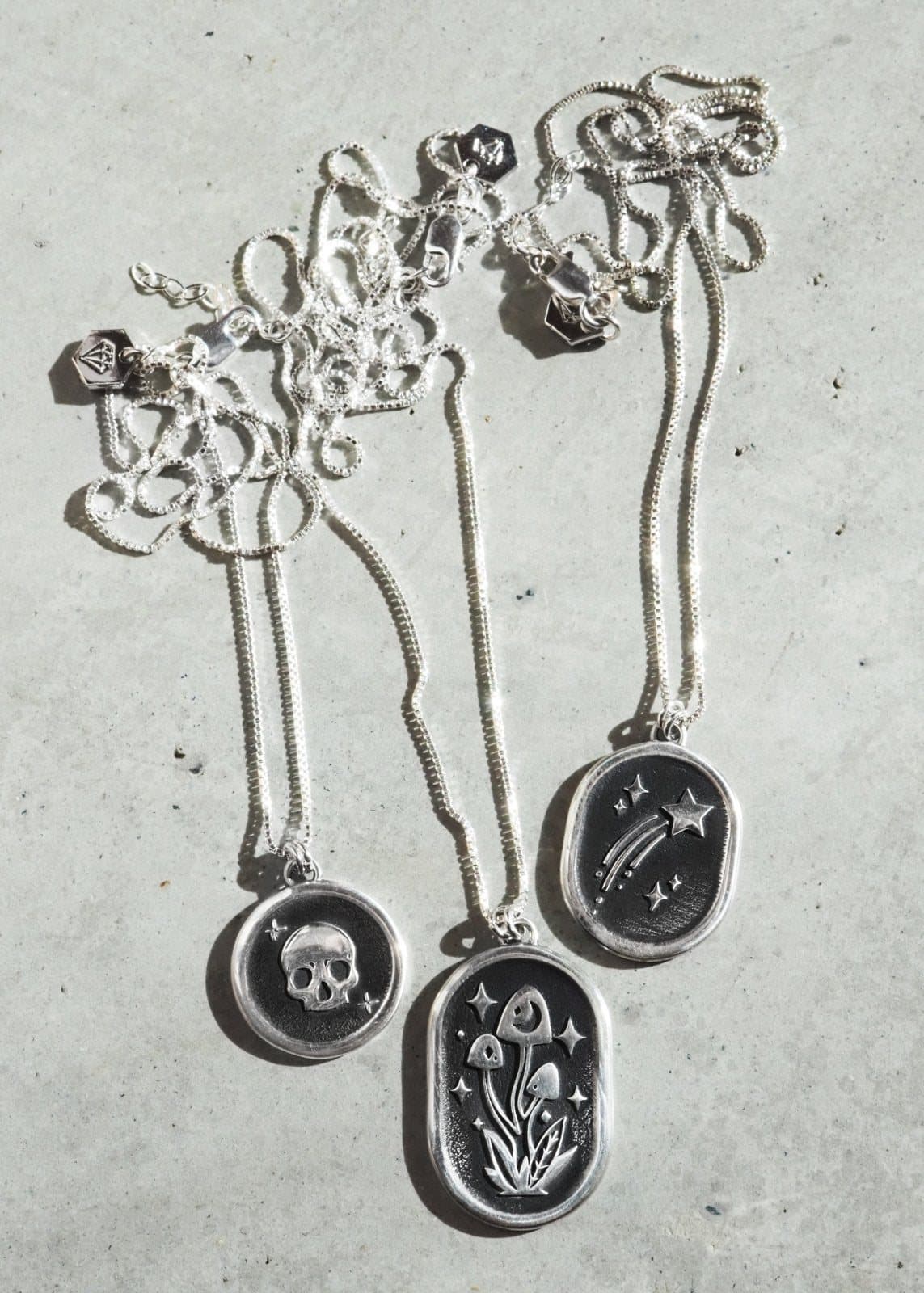 Three LanaBetty silver pendant necklaces with engraved designs laid on pale stone.