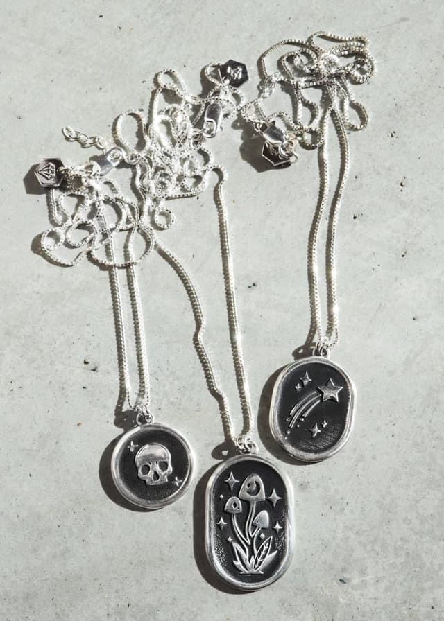 Three LanaBetty silver pendant necklaces with engraved designs laid on pale stone.