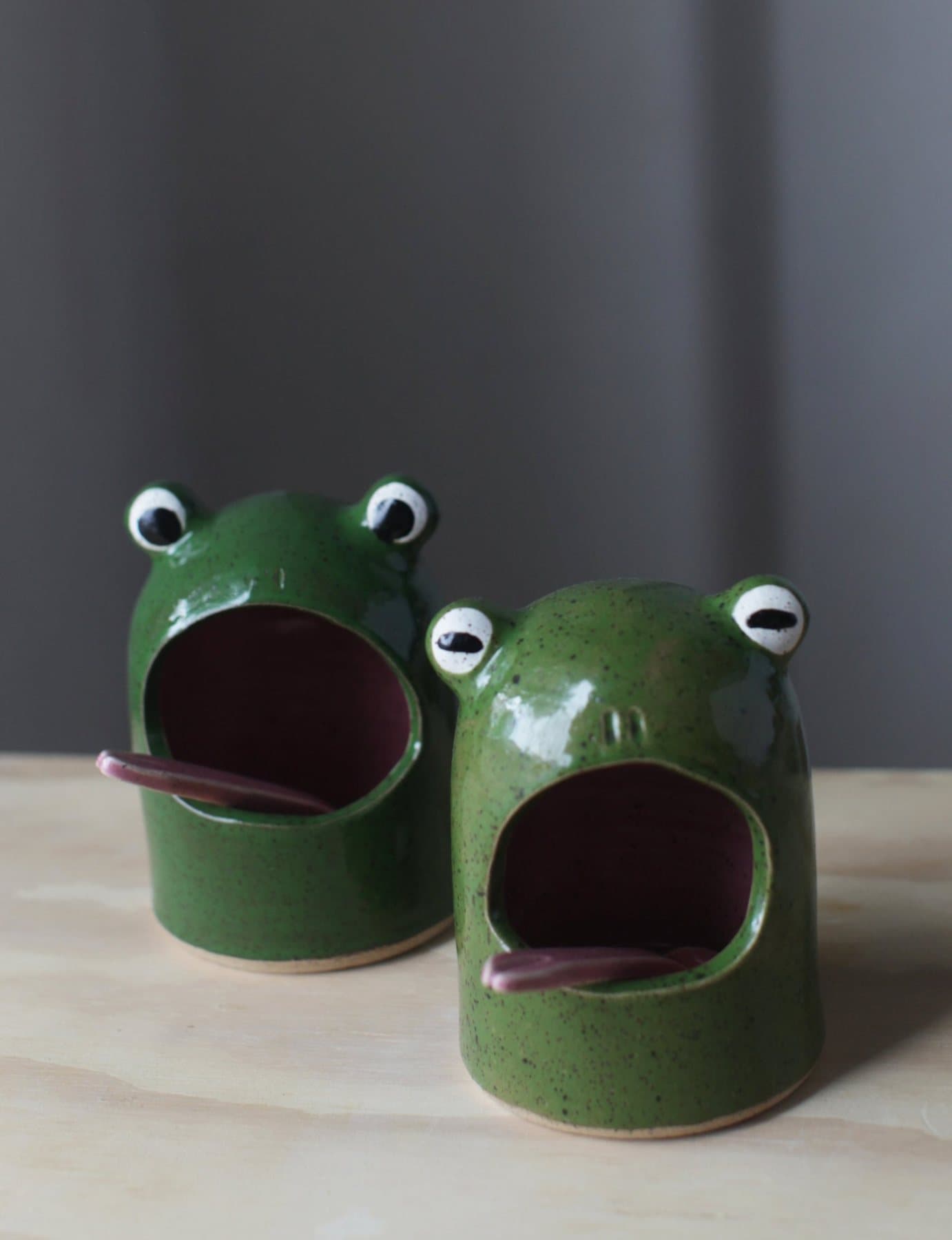 A pair of green frog salt cellars by Ohhelloleigh Ceramics with open mouths and pink tongues.