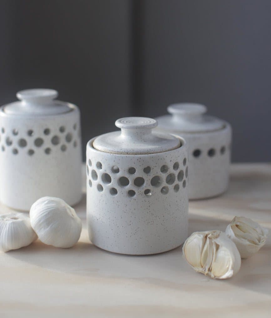 Three white Ohhelloleigh Ceramics garlic keepers with cut-out holes beside bulbs of garlic.