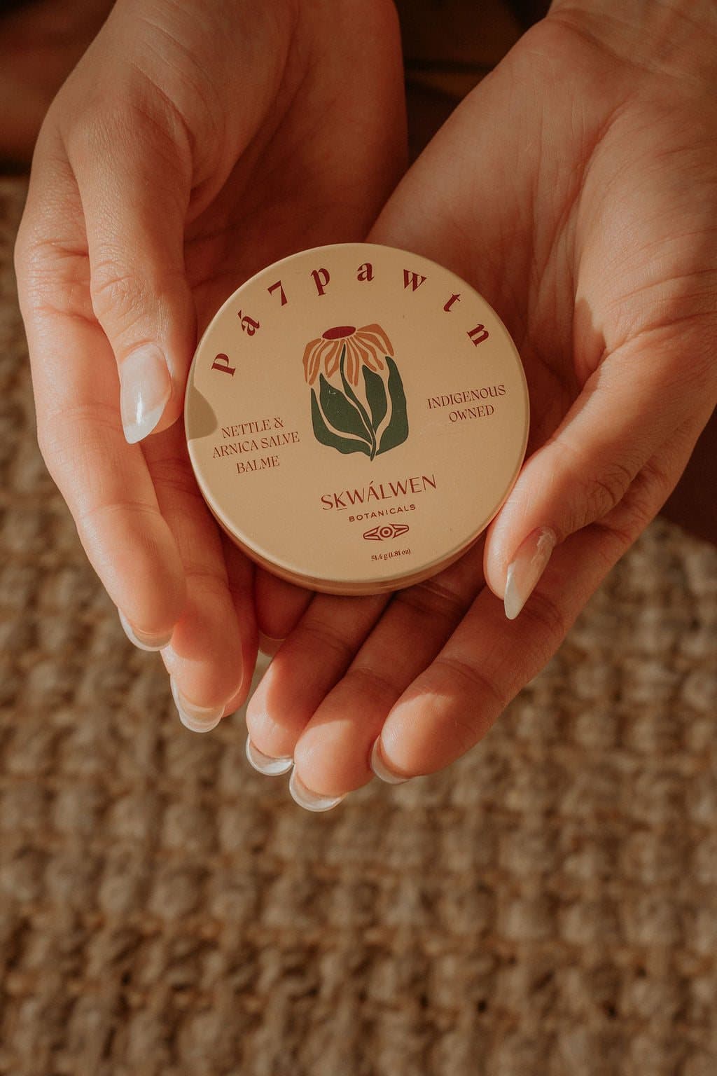 Two hands hold an open-lidded tin of Sḵwálwen Botanicals balm over a woven mat.