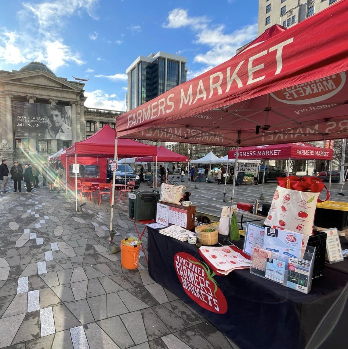 Red Farmers Markets tents and a stall table on the paved square at the Downtown Farmers Market.