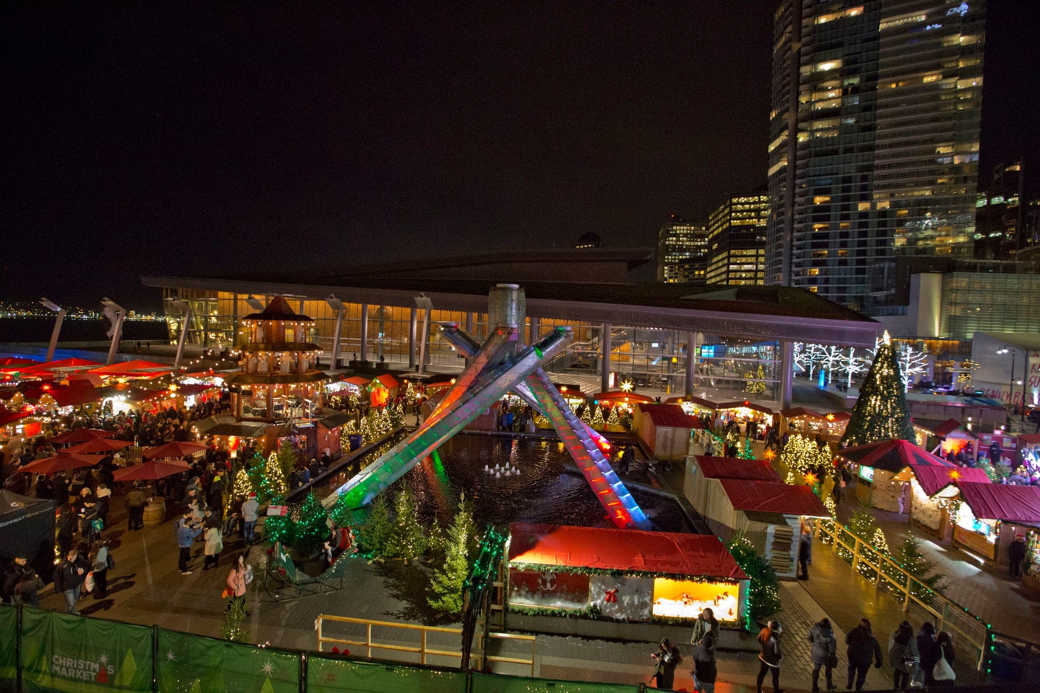 Wooden stalls and a lit Christmas pyramid fill Jack Poole Plaza at the Vancouver Christmas Market at night.