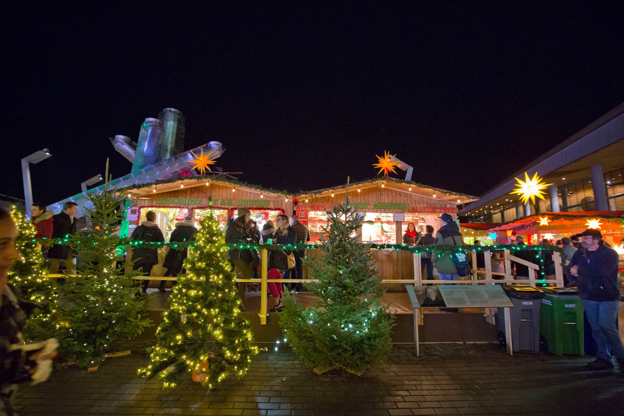 Lit trees and wooden stalls of the Vancouver Christmas Market stand below the Olympic Cauldron on Jack Poole Plaza.