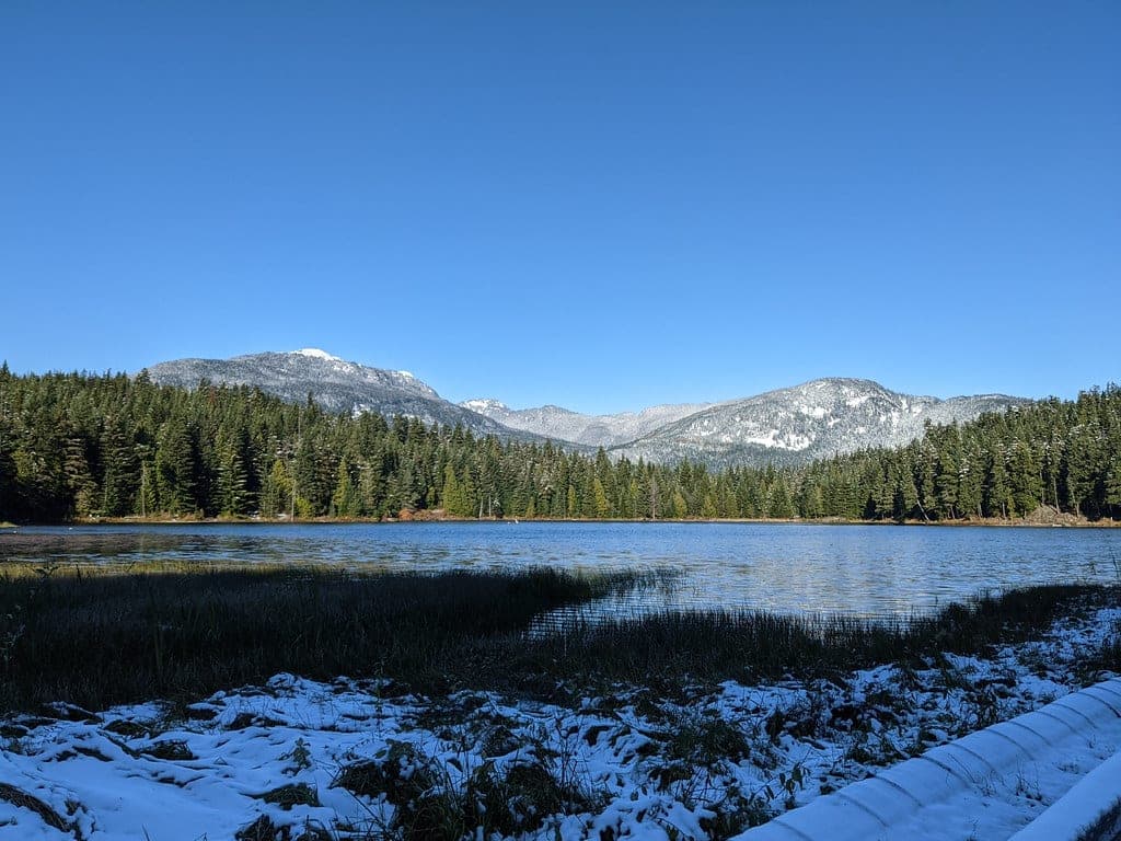 Early snow dusts the shore of Lost Lake in Whistler under a clear sky and snowy peaks.