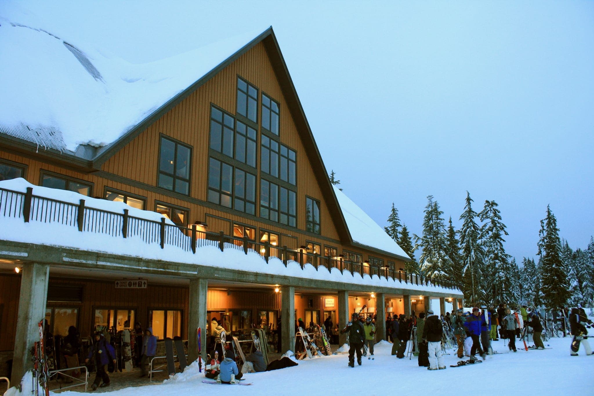 Skiers and snowboarders gather outside the Cypress Mountain day lodge at dusk with snow on the roof.