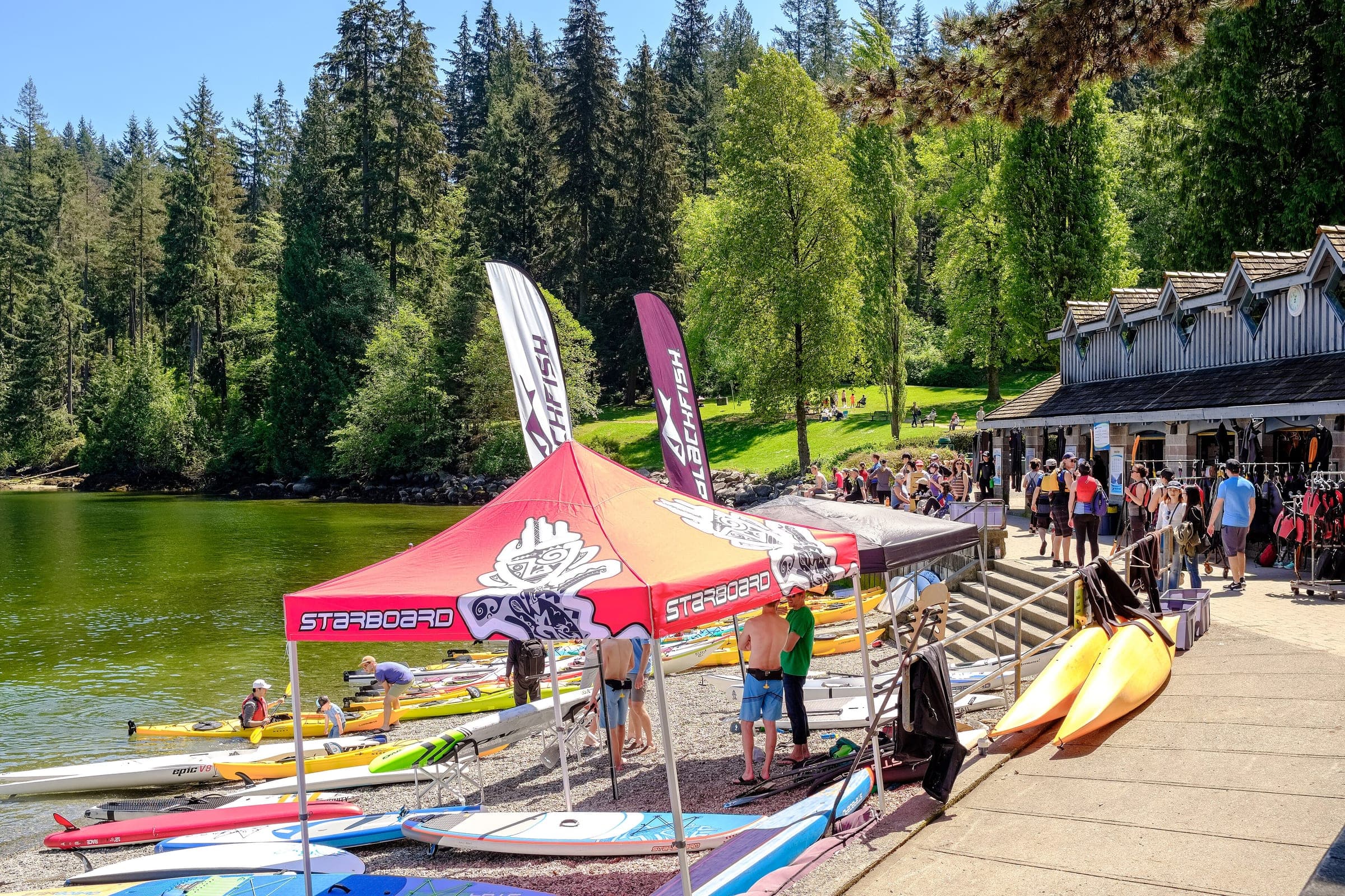 Paddlers and rental kayaks gather on the Deep Cove shore beside a boathouse in summer.