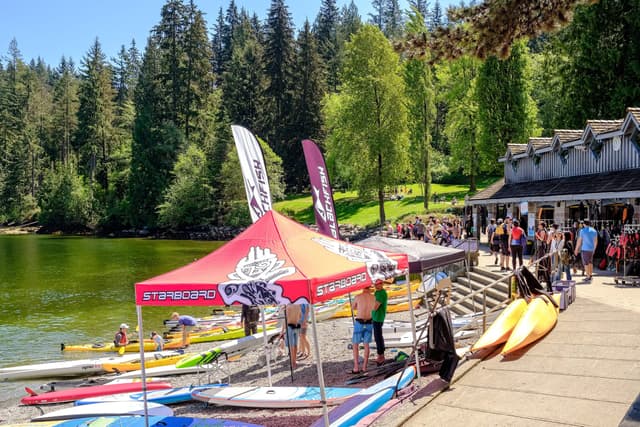 Paddlers and rental kayaks gather on the Deep Cove shore beside a boathouse in summer.