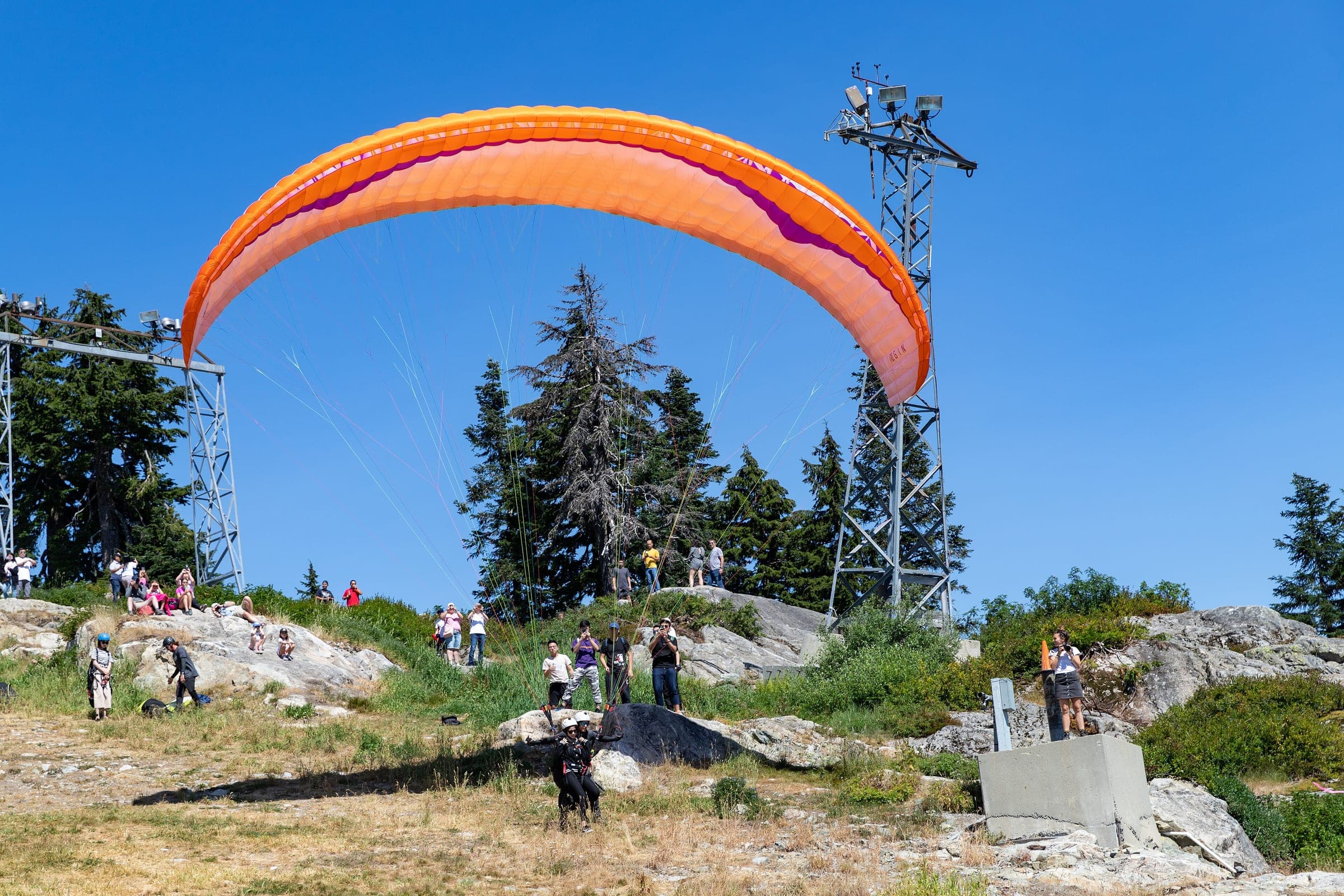 A paraglider inflates an orange wing at the Grouse Mountain launch while visitors watch from the rocks.