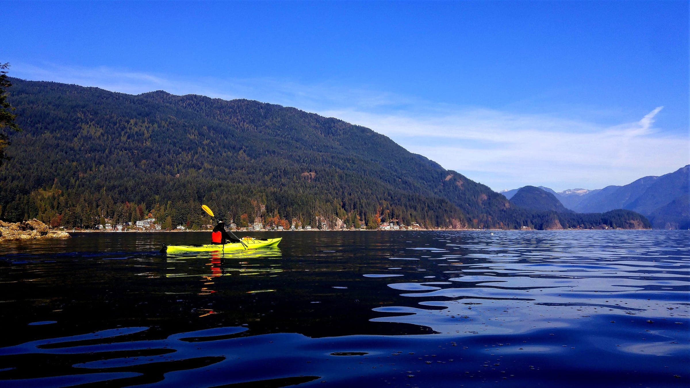 A kayaker paddles a yellow boat across calm water in Deep Cove below the forested ridge of Indian Arm.
