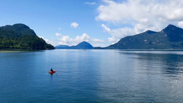 A kayaker crosses calm blue water off Porteau Cove with the forested peaks of Howe Sound all around.