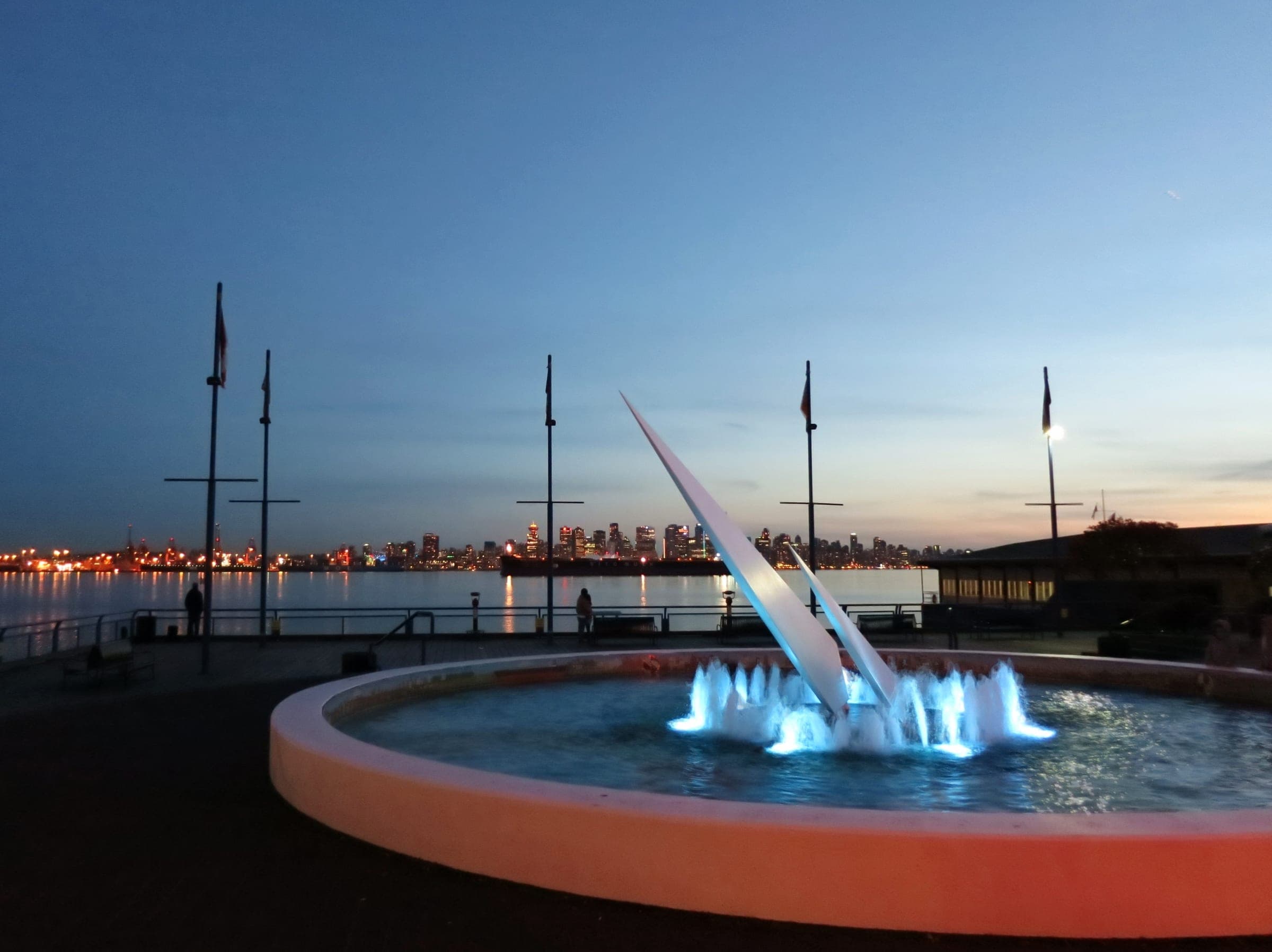 The fountain at Lonsdale Quay glows at dusk as the downtown Vancouver skyline lights up across the water.