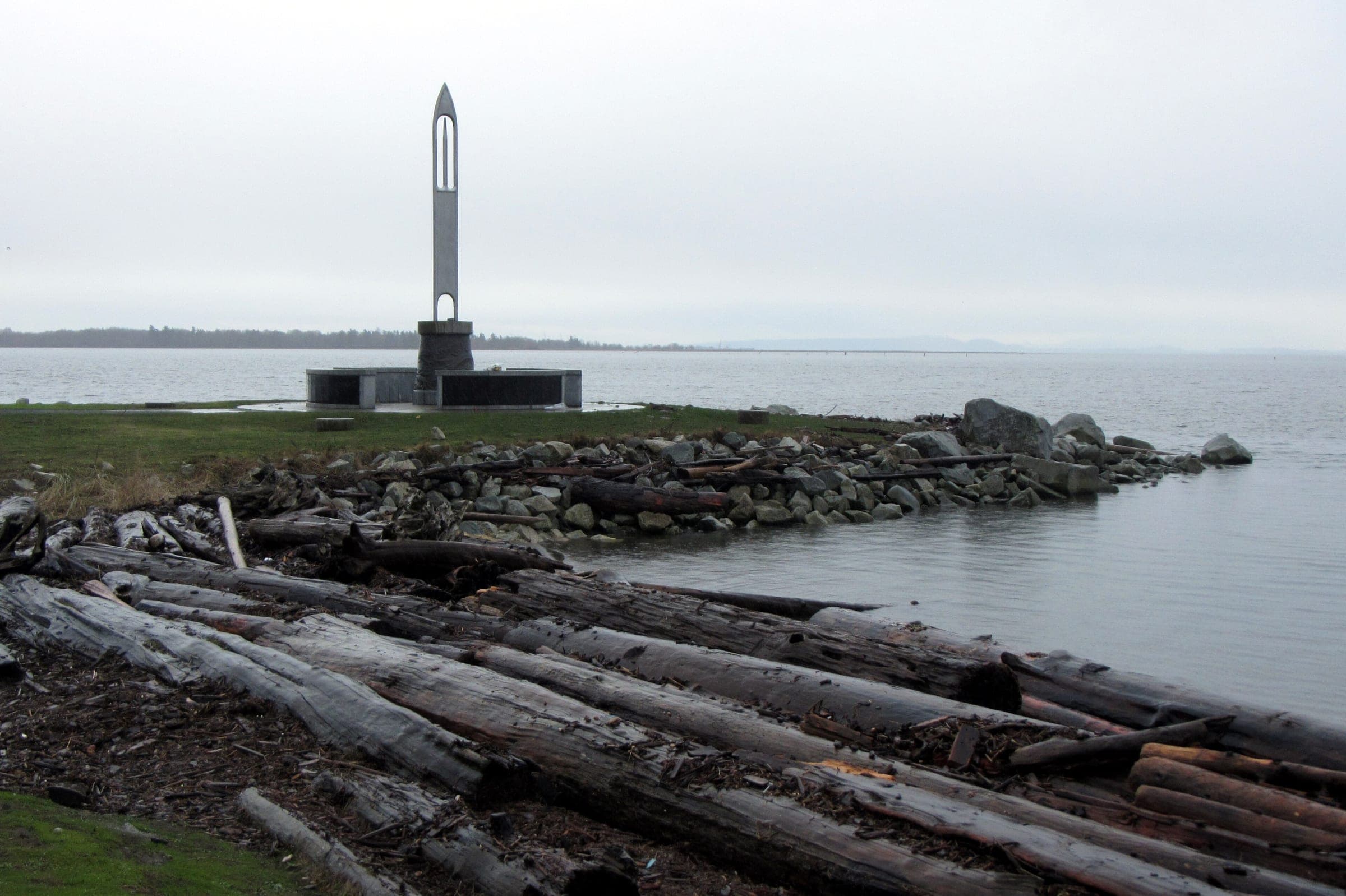 The Fisherman's Memorial needle stands above driftwood and boulders at Garry Point Park on a grey day.