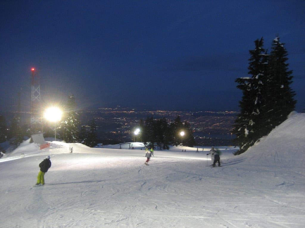 Skiers and snowboarders ride a floodlit Grouse Mountain run with the lights of Vancouver spread below.