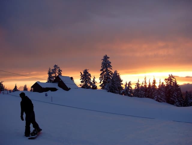 A snowboarder crosses the Grouse Mountain snow as the sun sets behind a cabin and snow-loaded firs.