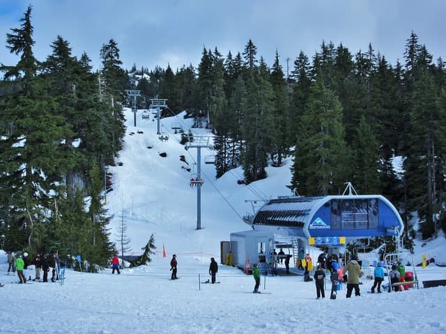 Skiers and snowboarders gather at the base of a Mount Seymour chairlift below snowy forest.