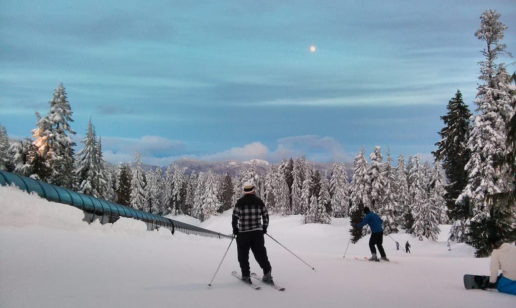 Skiers glide down a gentle Mount Seymour run at dusk as the moon rises over snow-loaded firs.