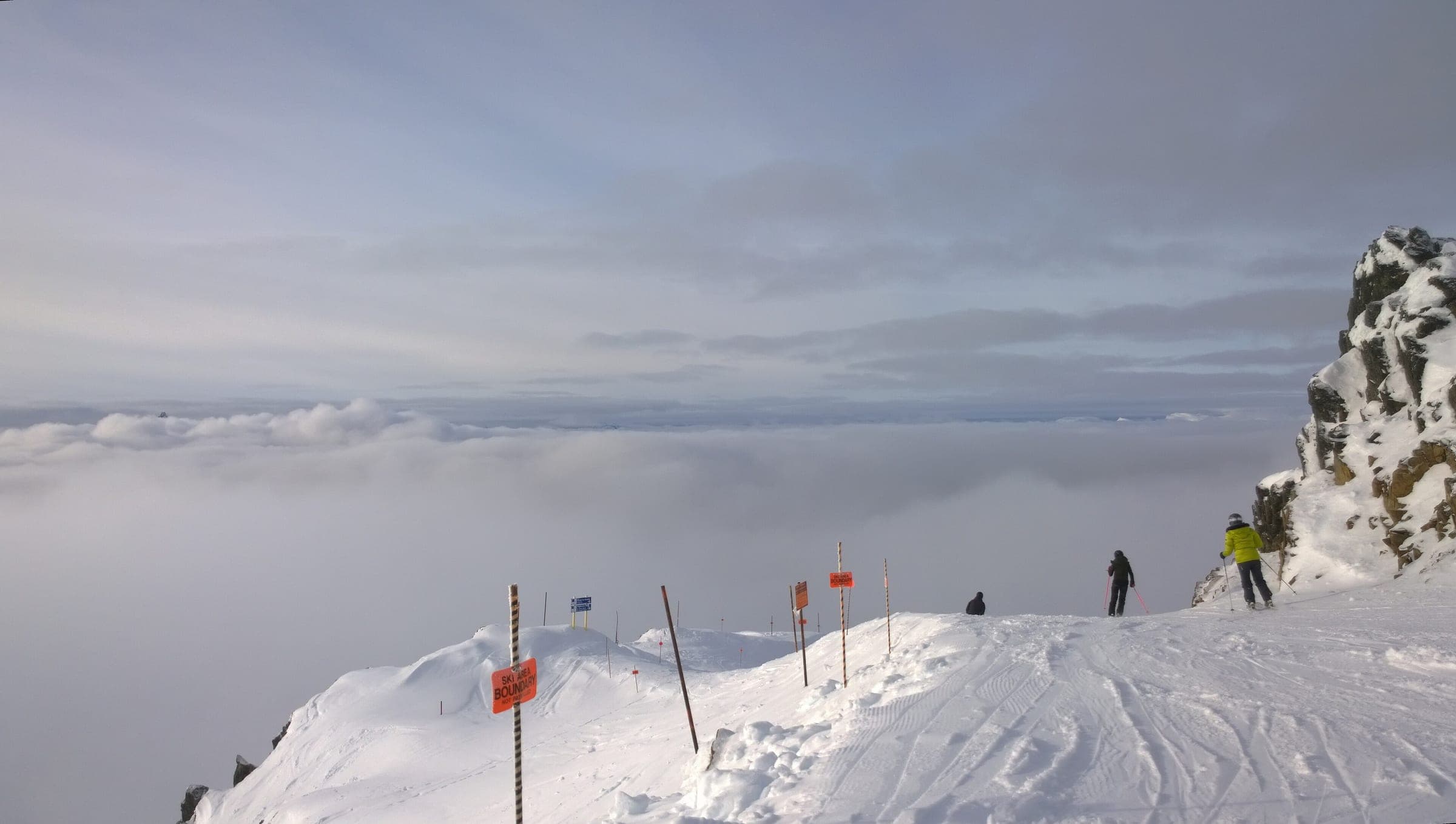Skiers stand on a Whistler Blackcomb ridge above a valley filled with cloud.