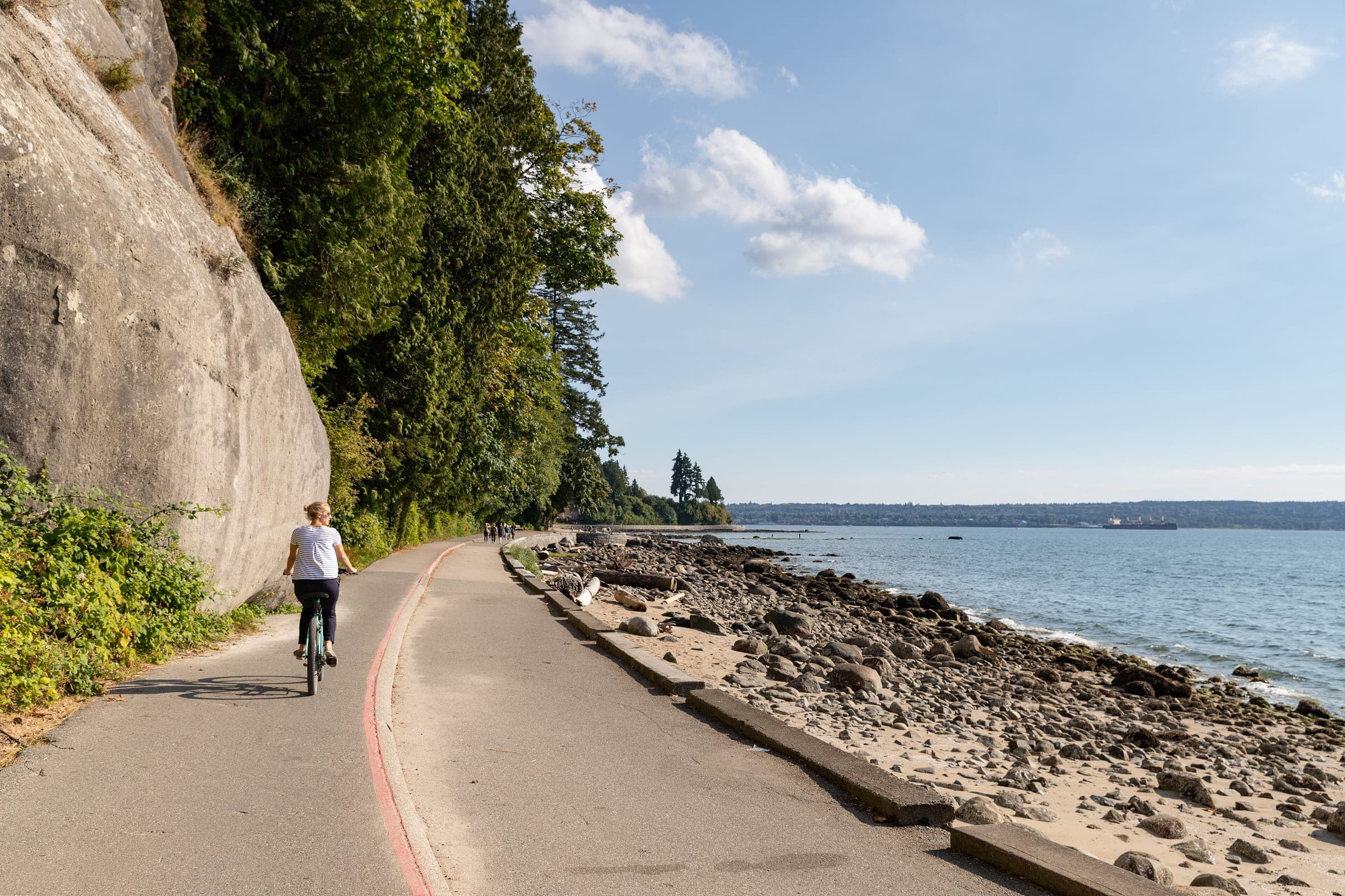 A cyclist rides the Stanley Park Seawall under a mossy cliff beside the rocky shore of English Bay.