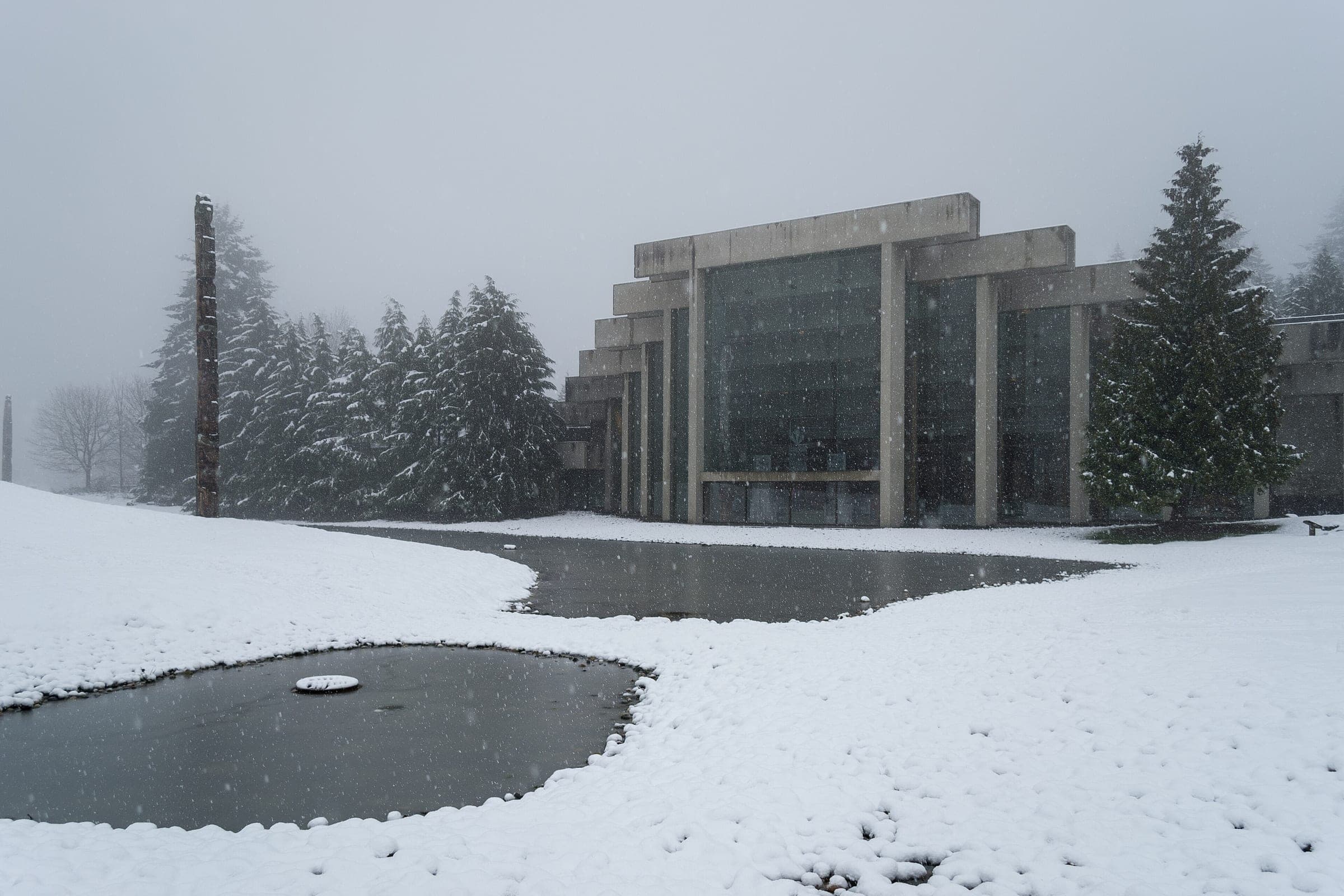 The Museum of Anthropology at UBC under fresh snow with its concrete and glass front beside the pond.