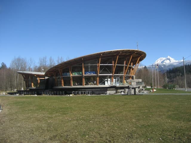 The timber-roofed Squamish Adventure Centre on Loggers Lane with snowy peaks beyond the field.
