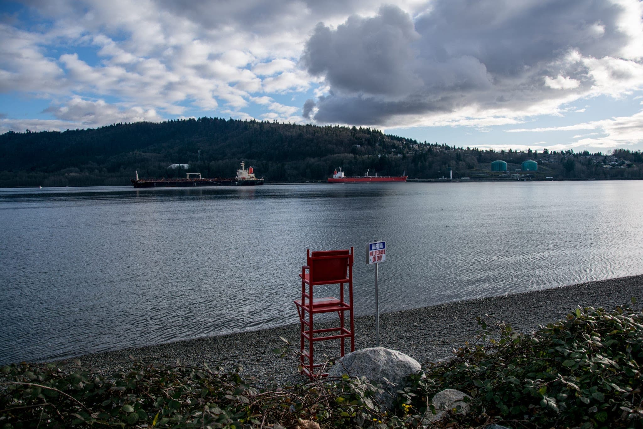A red lifeguard chair stands on the beach at Whey-ah-Wichen/Cates Park as tankers wait on Burrard Inlet.