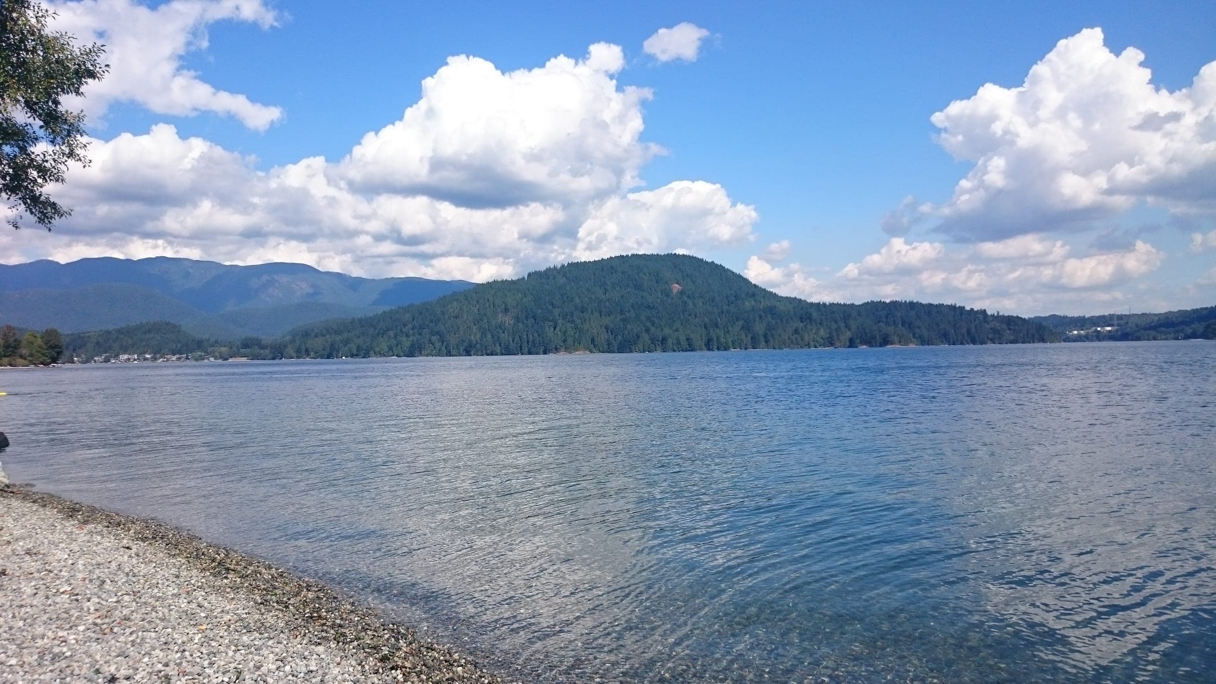 Calm water laps a pebble beach at Whey-ah-Wichen/Cates Park below forested hills across the inlet.