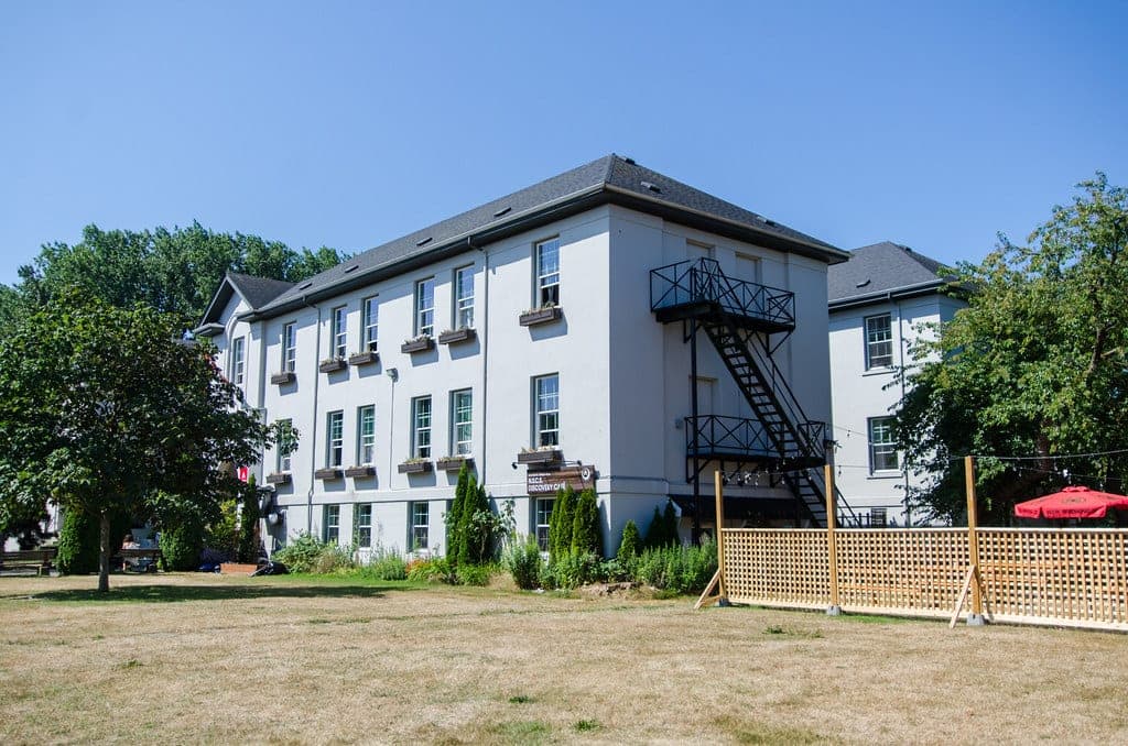 The white three-storey HI Vancouver Jericho Beach hostel stands among trees under a clear summer sky.