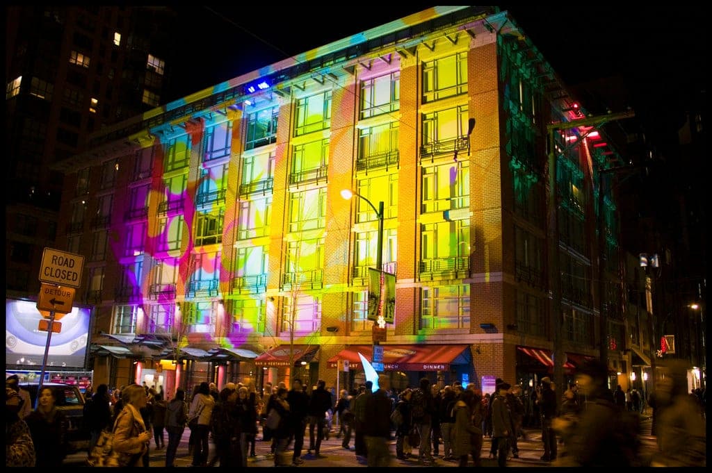 Crowds gather in front of Opus Hotel in Yaletown as coloured light plays across its facade.