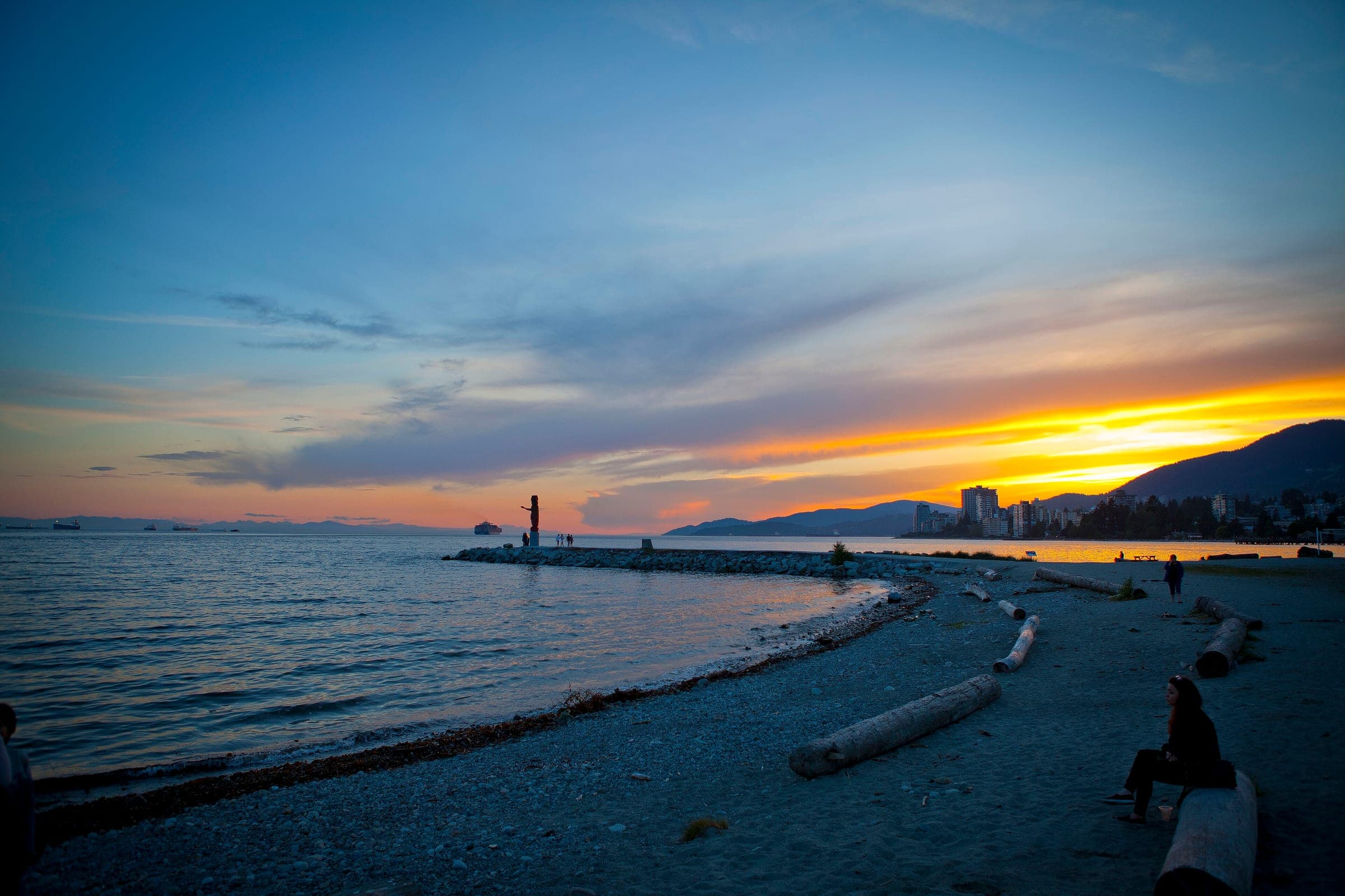The sun sets behind the West Vancouver hills as people sit among the driftwood on Ambleside Beach.