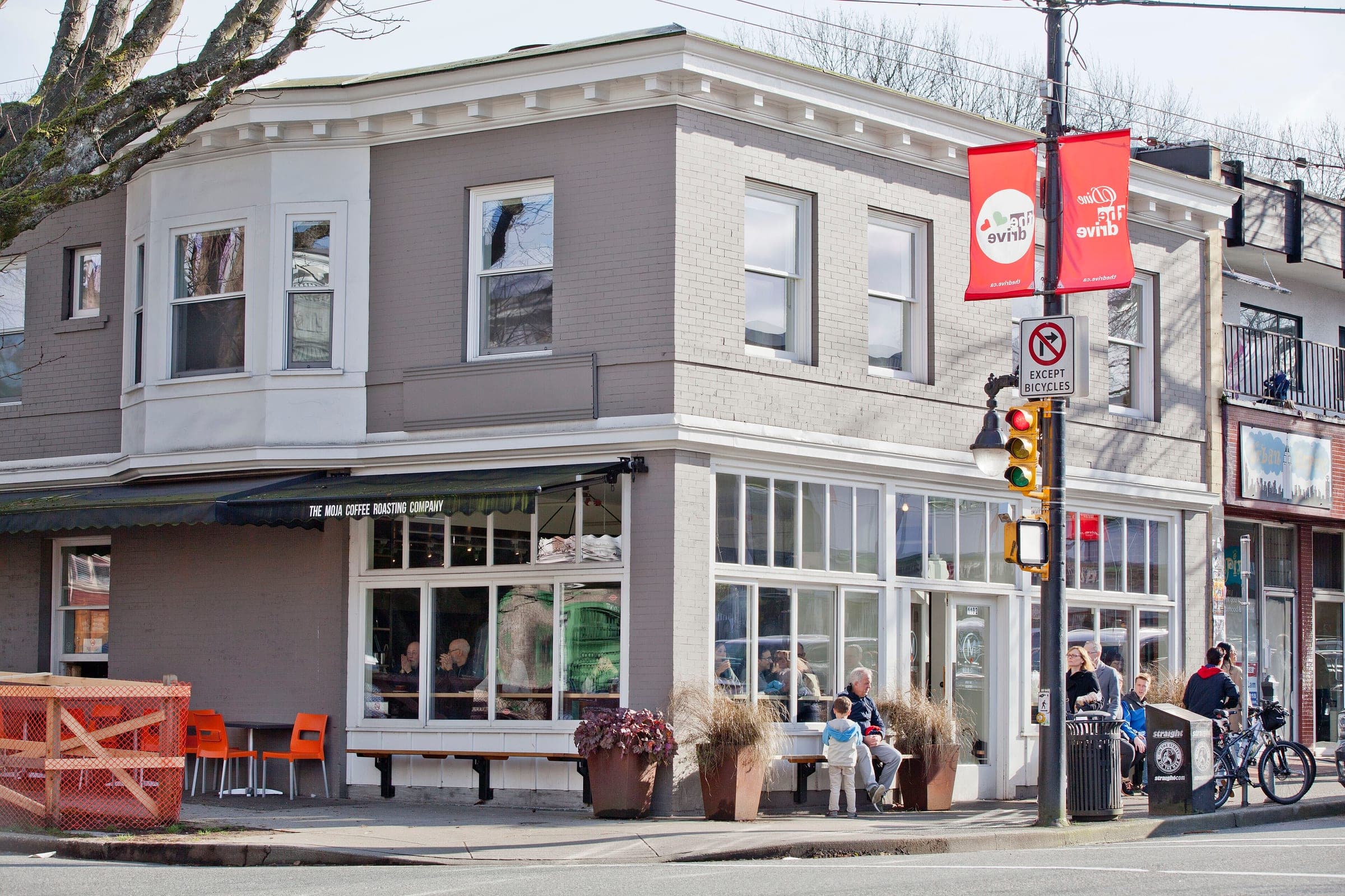 People sit outside a corner coffee roaster on Commercial Drive below its grey bay windows.