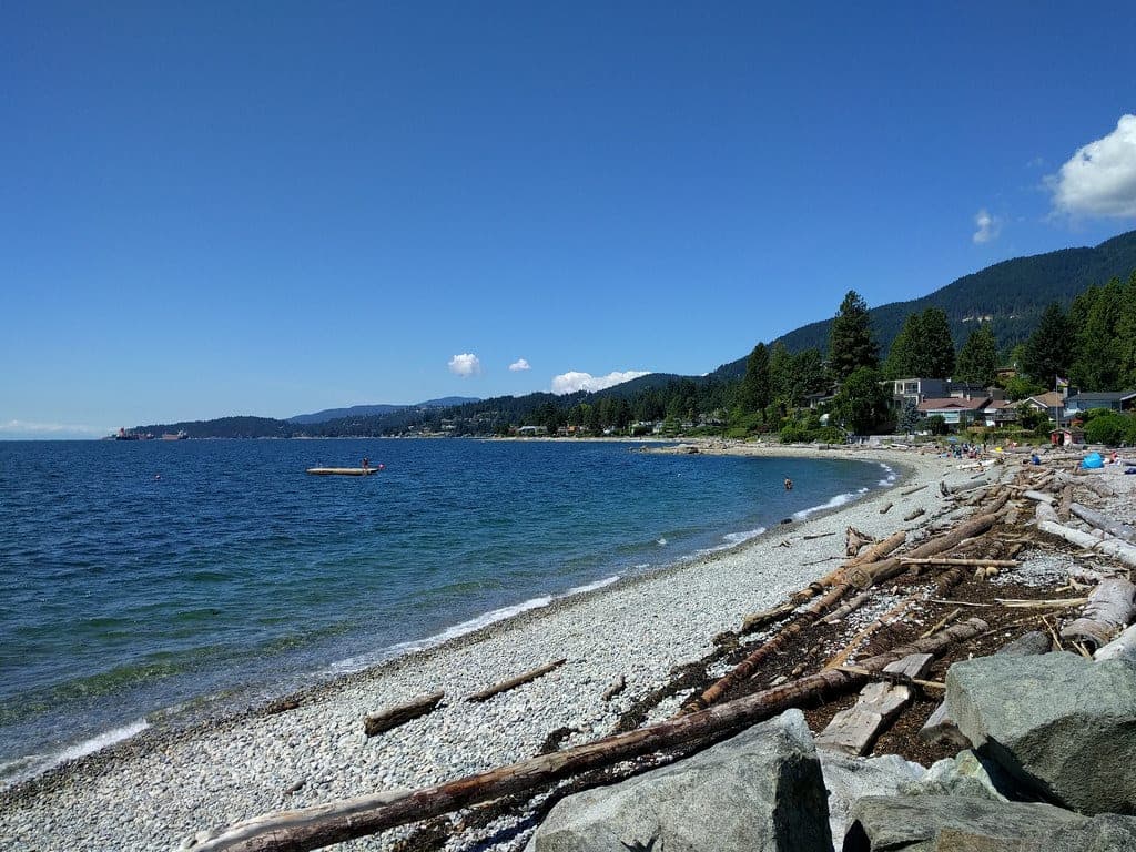 Driftwood lines the pebble shore of Dundarave Beach as the West Vancouver shoreline curves away under a clear sky.
