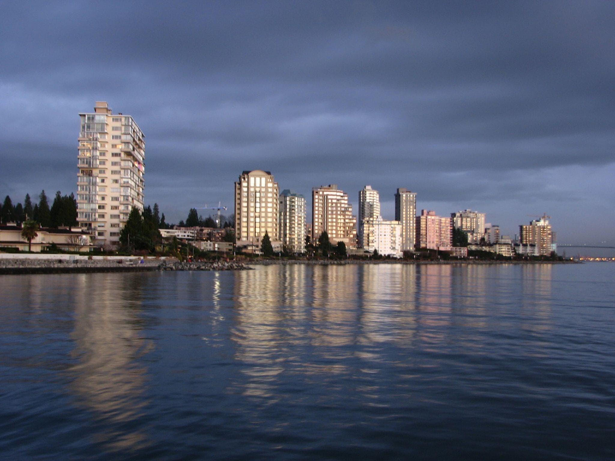 Evening light catches the apartment towers along the Dundarave shore under heavy grey cloud.