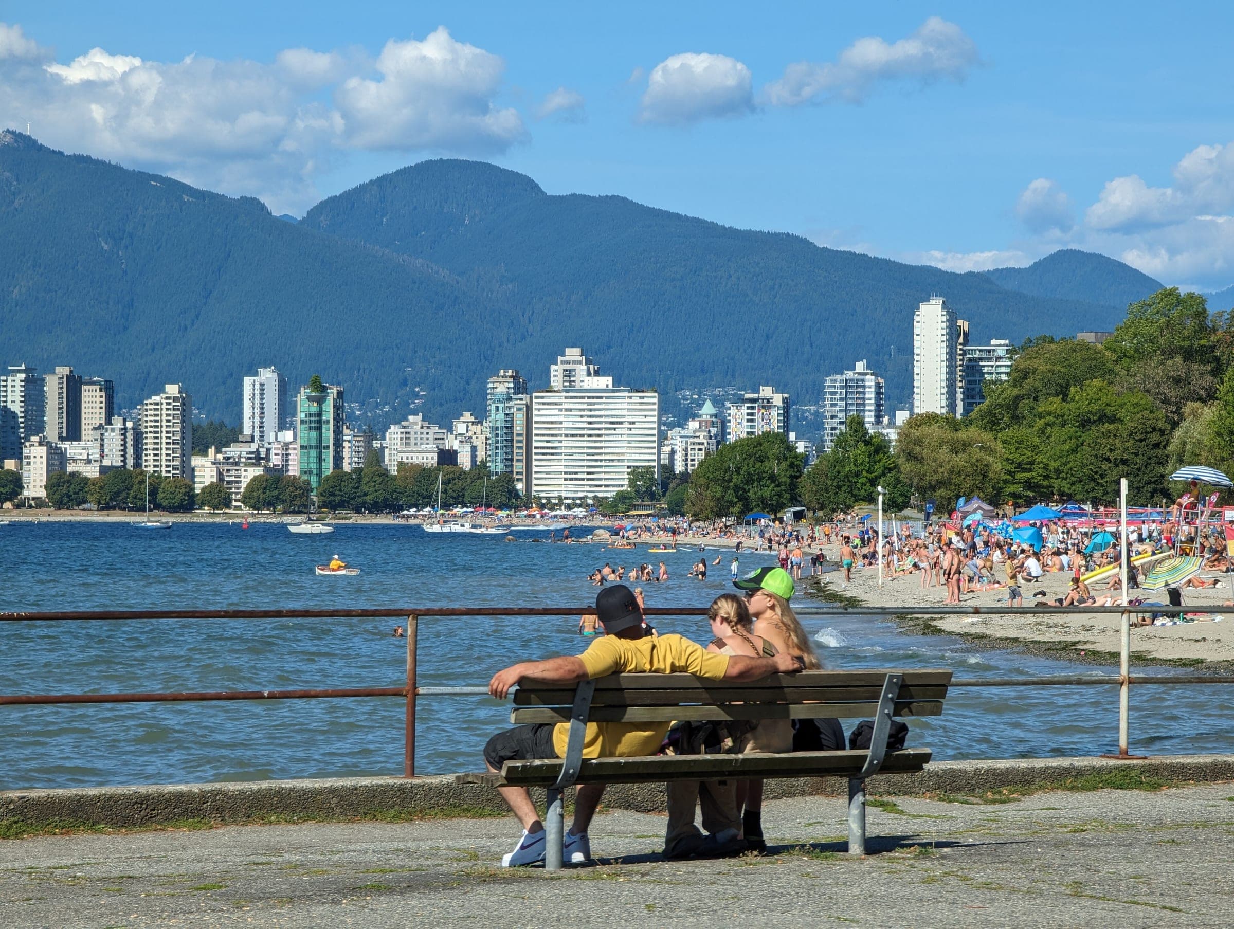 A couple sits on a bench above a crowded Kitsilano Beach facing the West End towers and the North Shore mountains.
